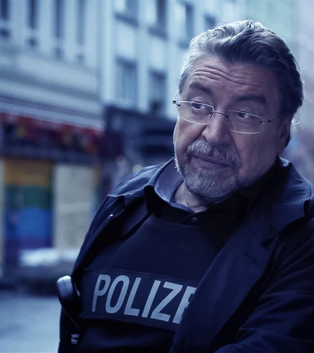 Ein Mann in Polizeiuniform (Armin Rohde) steht auf einer Straße und runzelt nachdenklich die Stirn. Im verschwommenen Hintergrund sieht man Häuserfassaden.