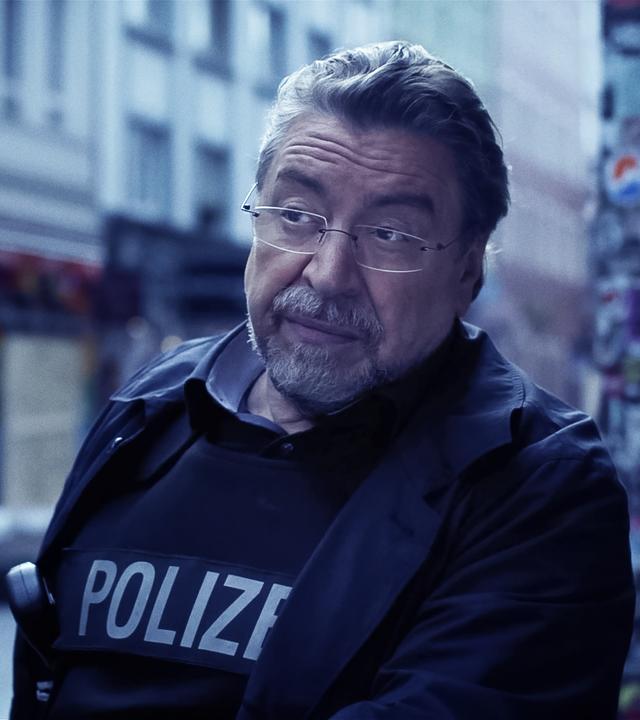 Ein Mann in Polizeiuniform (Armin Rohde) steht auf einer Straße und runzelt nachdenklich die Stirn. Im verschwommenen Hintergrund sieht man Häuserfassaden.