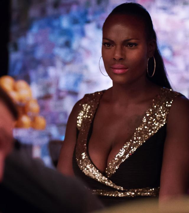 Eine Frau im Glitzerkleid schaut verärgert/ verwirrt auf einen Mann, der unscharf im Vordergrund zu erkennen ist. Der Mann lehnt an einem Geländer o. dergl.