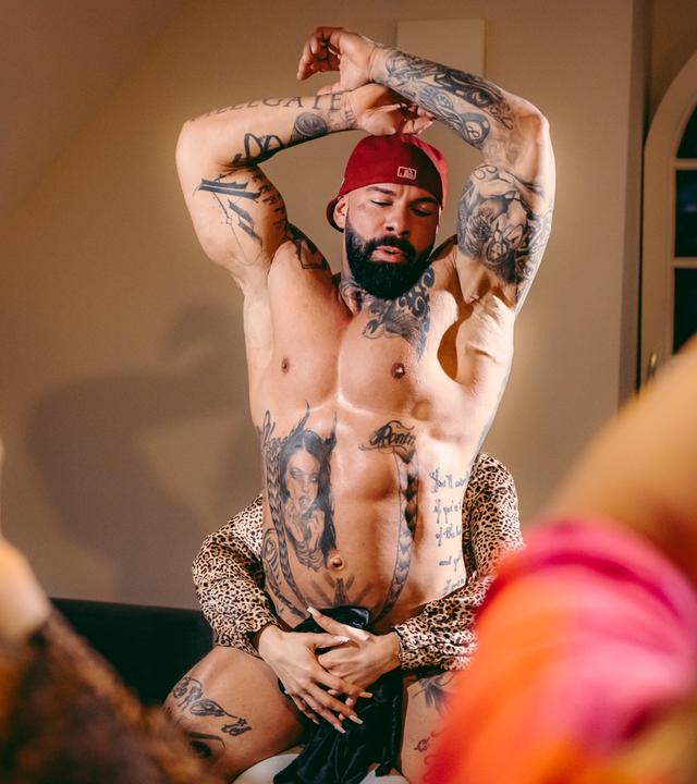 Leif steht bei seiner Performance in einem Raum. Er ist bis auf ein Tuch im Intimbereich nackt und hat die Arme gehoben. Von hinten hat eine Frau ihre Arme um seinen Bauch gelegt. An den Seiten im Anschnitt sind verschwommen die Rückenansichten von  Zuschauenden zu sehen.
