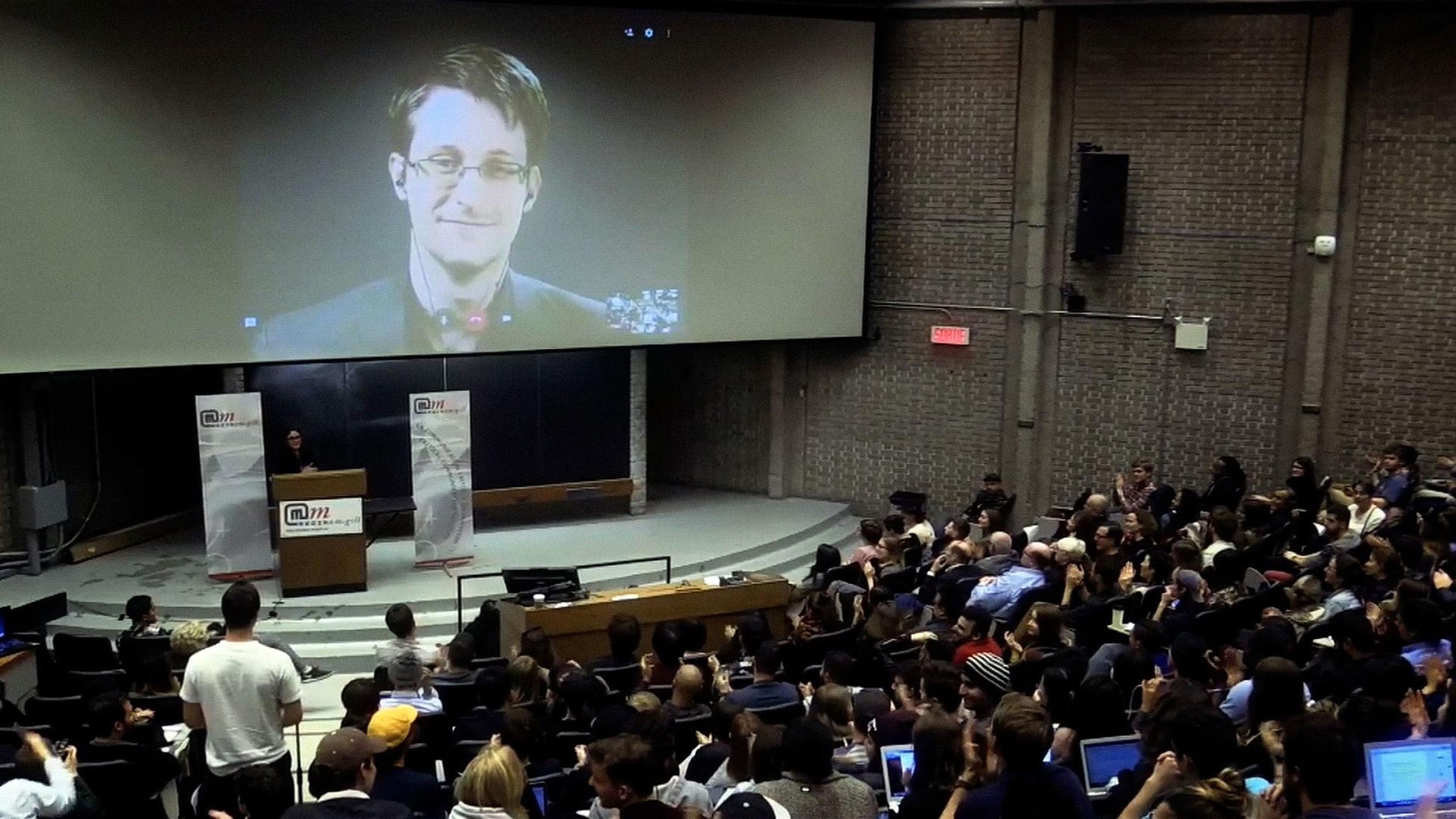 "Nackt im Netz - Hacker gegen Überwachung": Edward Snowden groß auf einer Leinwand, in einem Saal sitzen Menschen vor Bildschirmen.