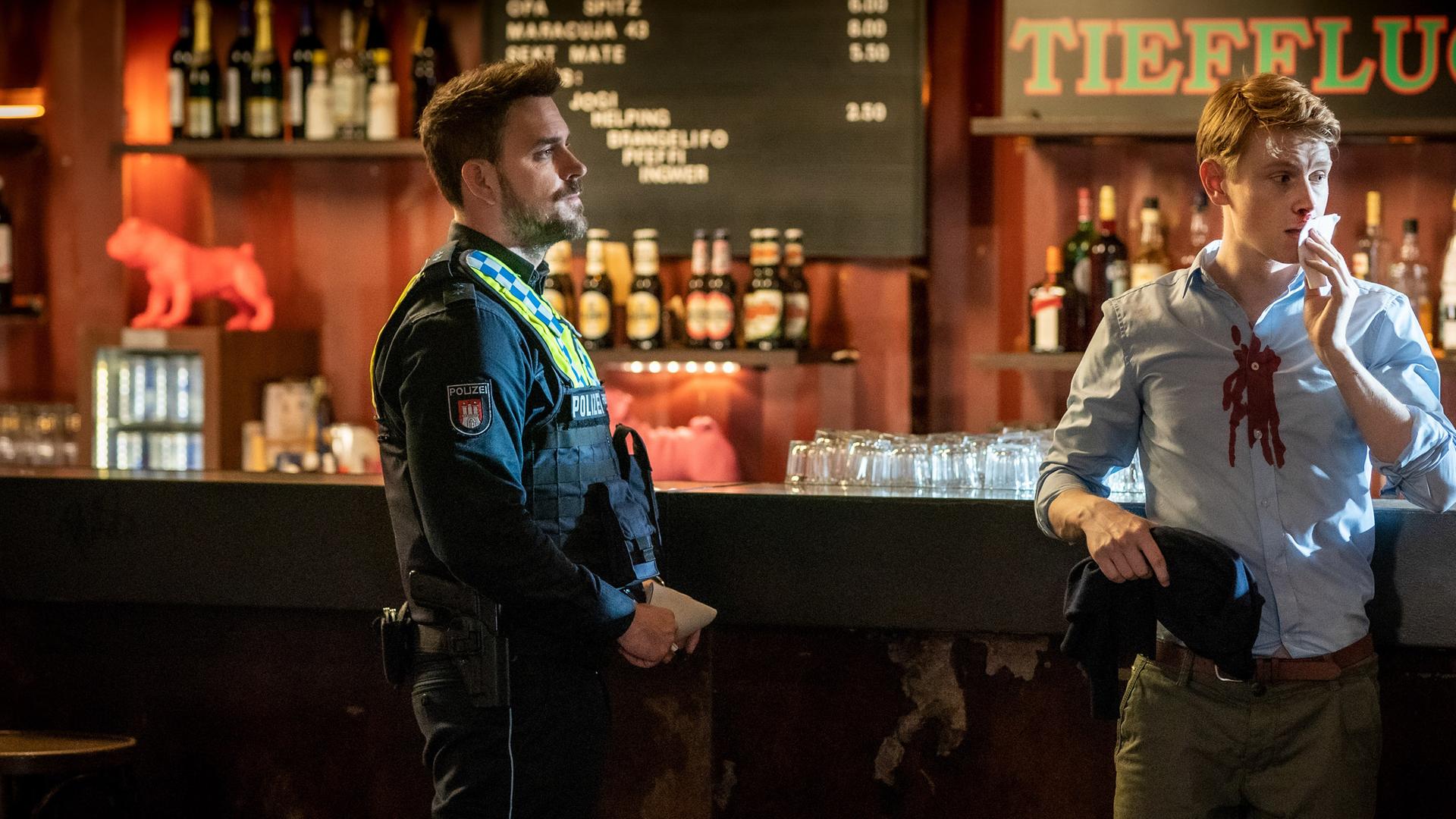 Der Polizist Kris Freiberg (Marc Barthel) steht in einem Club vor der Theke. Neben ihm steht Nikolas Breuer (Lukas Zumbrock). Er lehnt mit dem Rücken an der Theke. Nikolas' Hemd hat Blutflecken. Er tupft sich mit einer Serviette die blutende Nase. Er blickt zur Seite. Kris sieht Nikolas an.