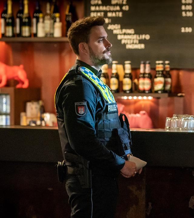 Der Polizist Kris Freiberg (Marc Barthel) steht in einem Club vor der Theke. Neben ihm steht Nikolas Breuer (Lukas Zumbrock). Er lehnt mit dem Rücken an der Theke. Nikolas' Hemd hat Blutflecken. Er tupft sich mit einer Serviette die blutende Nase. Er blickt zur Seite. Kris sieht Nikolas an.