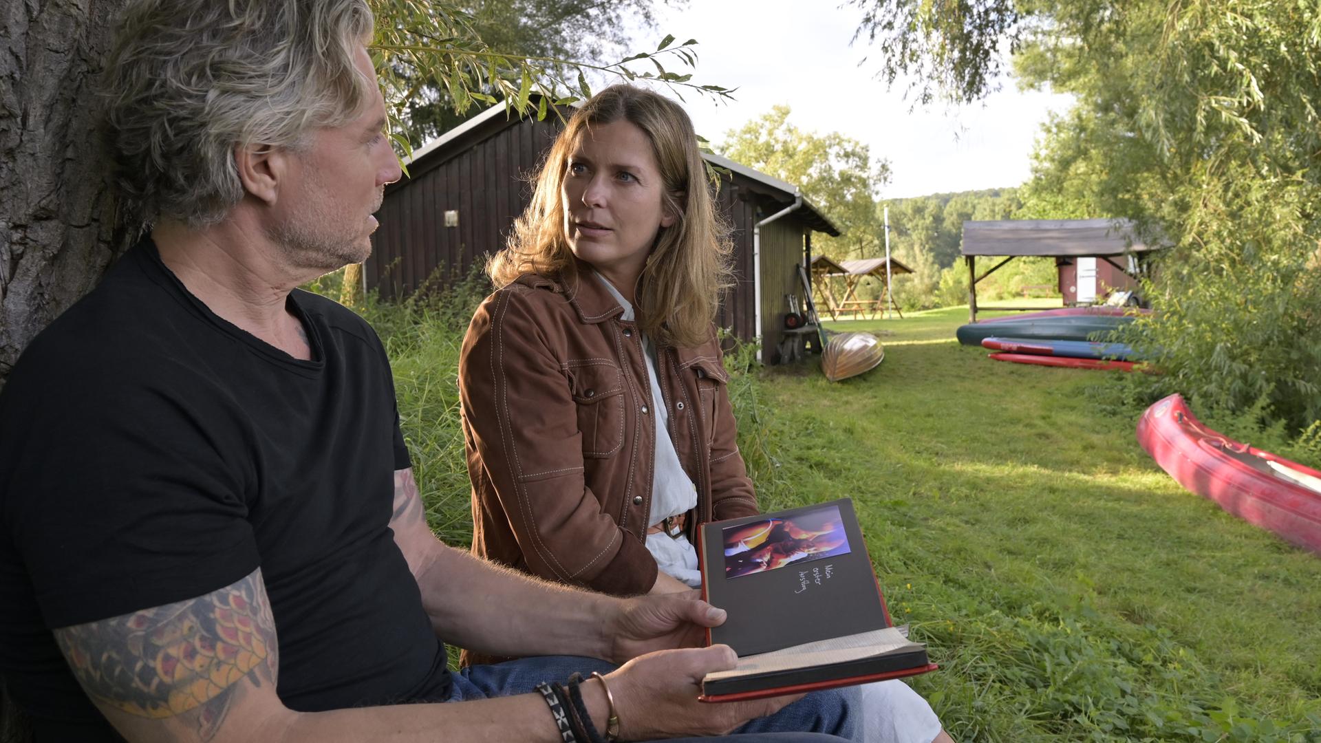 Katharina (Valerie Niehaus) sitzt mit Juri (Dirk Borchardt) im Garten unter einem Baum. Juri hält ein Fotoalbum in den Händen.