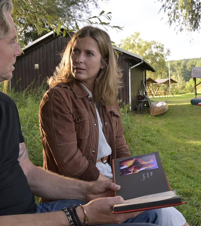 Katharina (Valerie Niehaus) sitzt mit Juri (Dirk Borchardt) im Garten unter einem Baum. Juri hält ein Fotoalbum in den Händen.