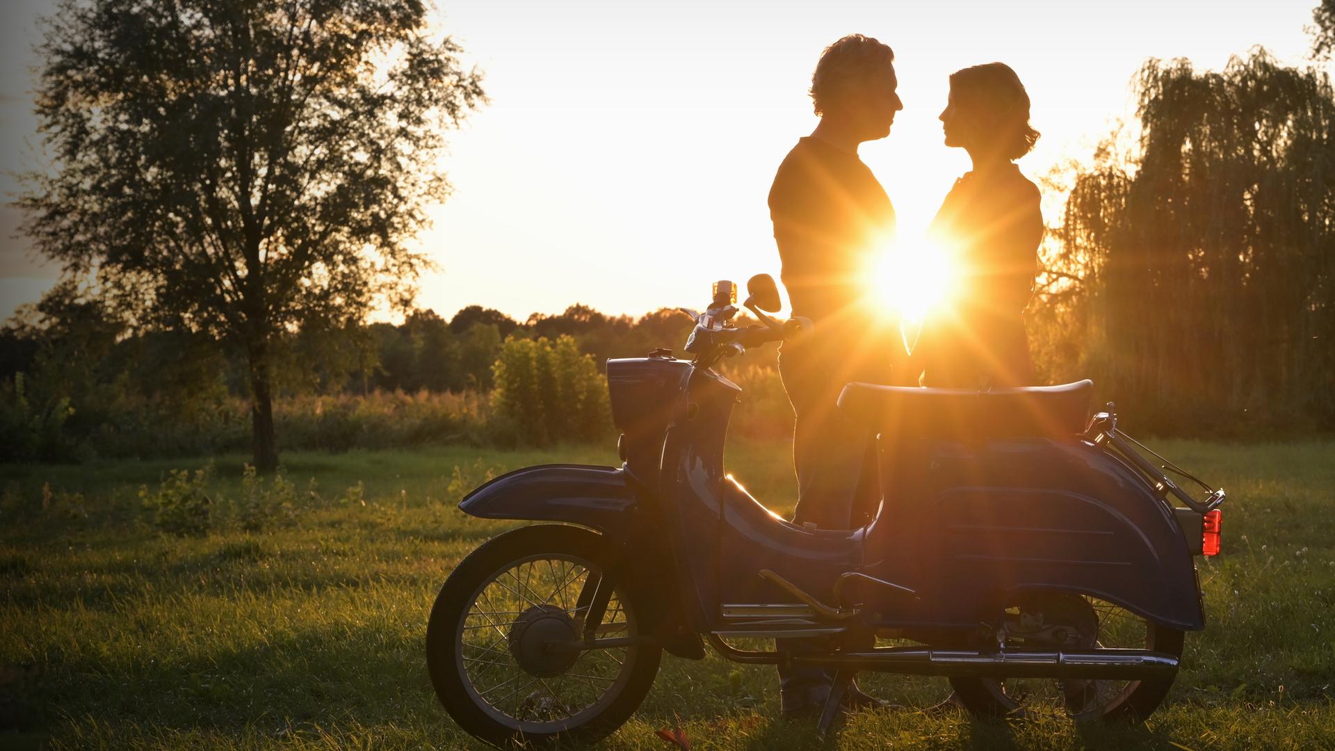 "Nächste Ausfahrt Glück - Familienbesuch": Juri Hoffmann (Dirk Borchardt) und Katharina Wegener (Valerie Niehaus) stehen im Gegenlicht eines sommerlichen Sonnenuntergangs, hinter ihnen Bäume und eine Wiese. Vor ihnen steht ein Moped der DDR-Marke "Schwalbe".