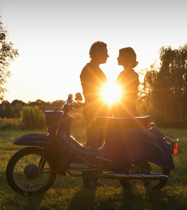 "Nächste Ausfahrt Glück - Familienbesuch": Juri Hoffmann (Dirk Borchardt) und Katharina Wegener (Valerie Niehaus) stehen im Gegenlicht eines sommerlichen Sonnenuntergangs, hinter ihnen Bäume und eine Wiese. Vor ihnen steht ein Moped der DDR-Marke "Schwalbe".