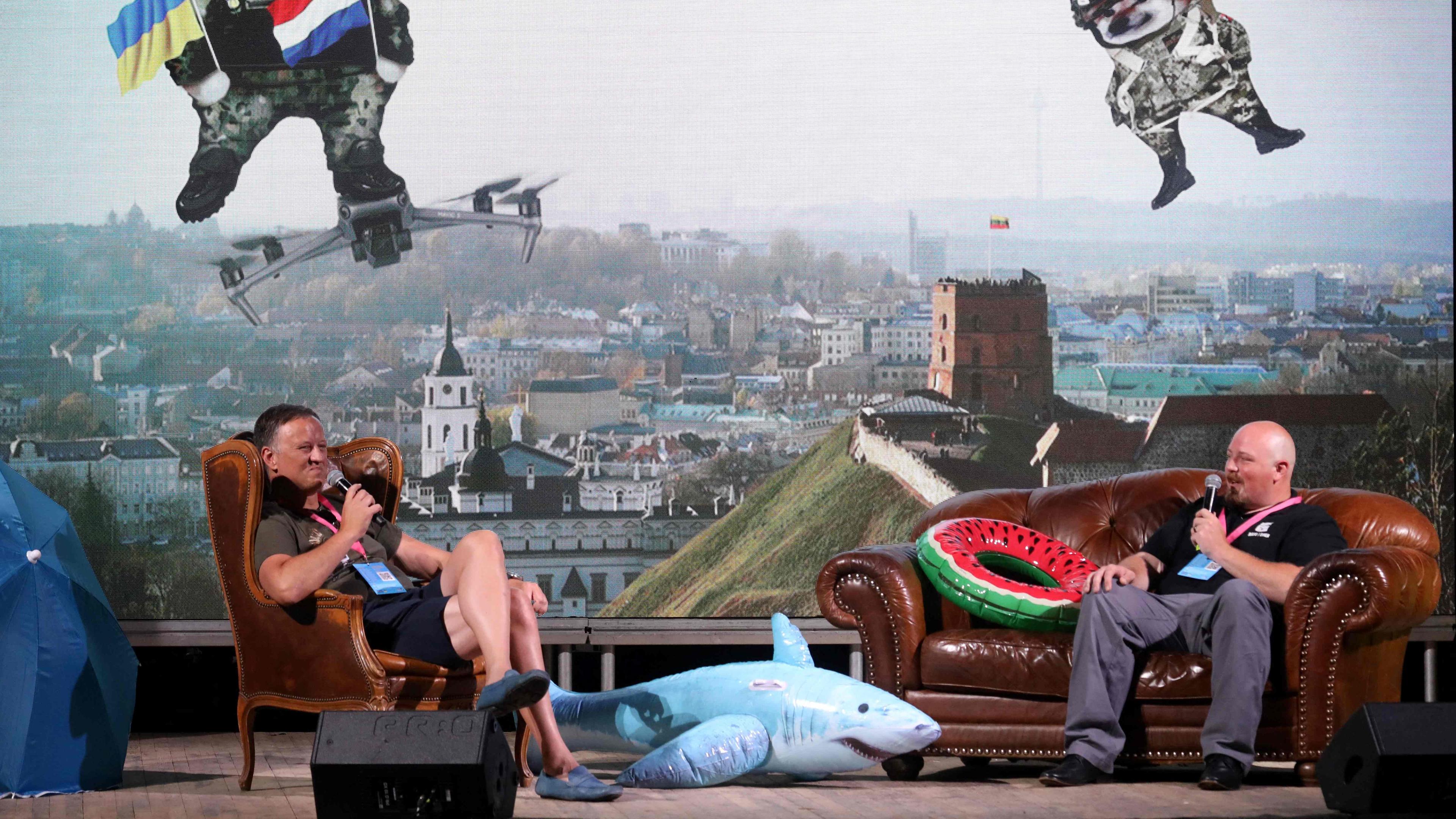 Zwei Männer sitzen auf Sesseln auf einer Bühne und unterhalten sich mit Mikros in der Hand. Im Hintergrund ist ein buntes Bild zu sehen, auf dem animierte Hunde im Himmel schweben.