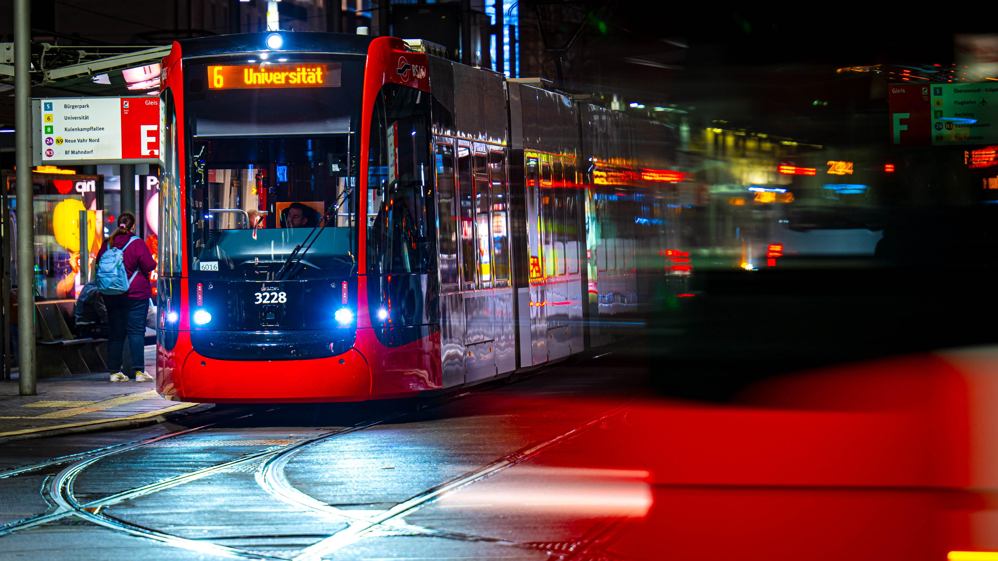 Im hintergrund hält eine rote Straßenbahn bei Nacht an einer Haltestelle. Im Vordergrund fährt ein roter Bus vorbei.