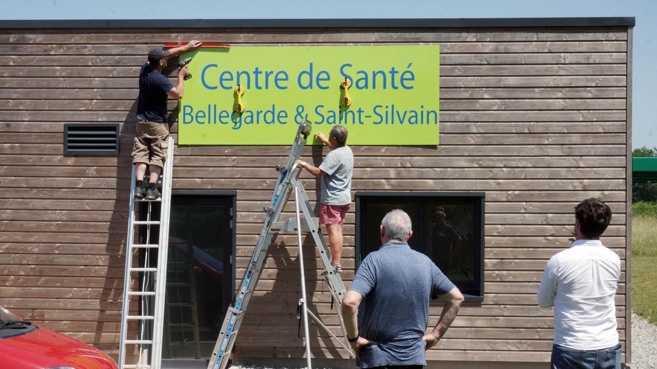An einem Holzhaus wird ein Schild mit der Aufschrift Centre de Santé, Bellegarde&Saint-Silvain angebracht - das zweite Gesundheitszentrum des Projekts.