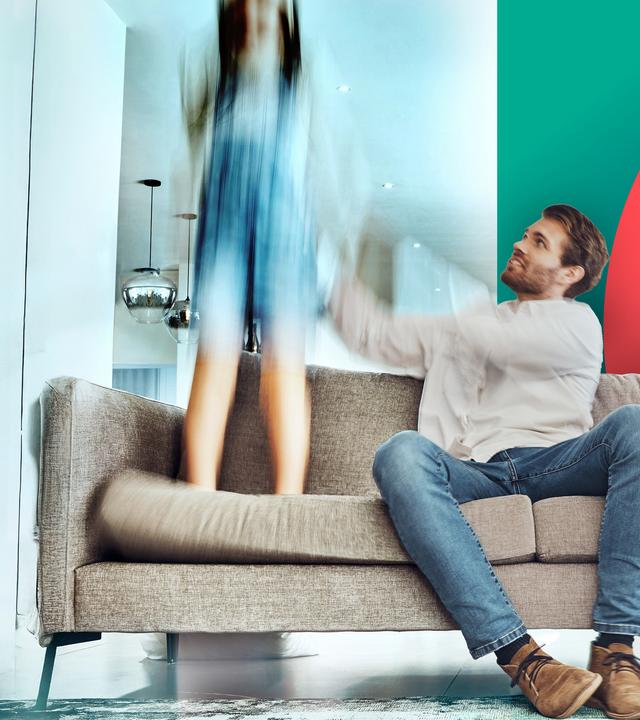 Ein junger Vater und eine junge Mutter sitzen auf einem Sofa. Die Mutter stützt den Kopf in die Hand und sieht erschöpft aus, der Vater reagiert entnervt mit einem Kind, das auf dem Sofa herumspringt. 