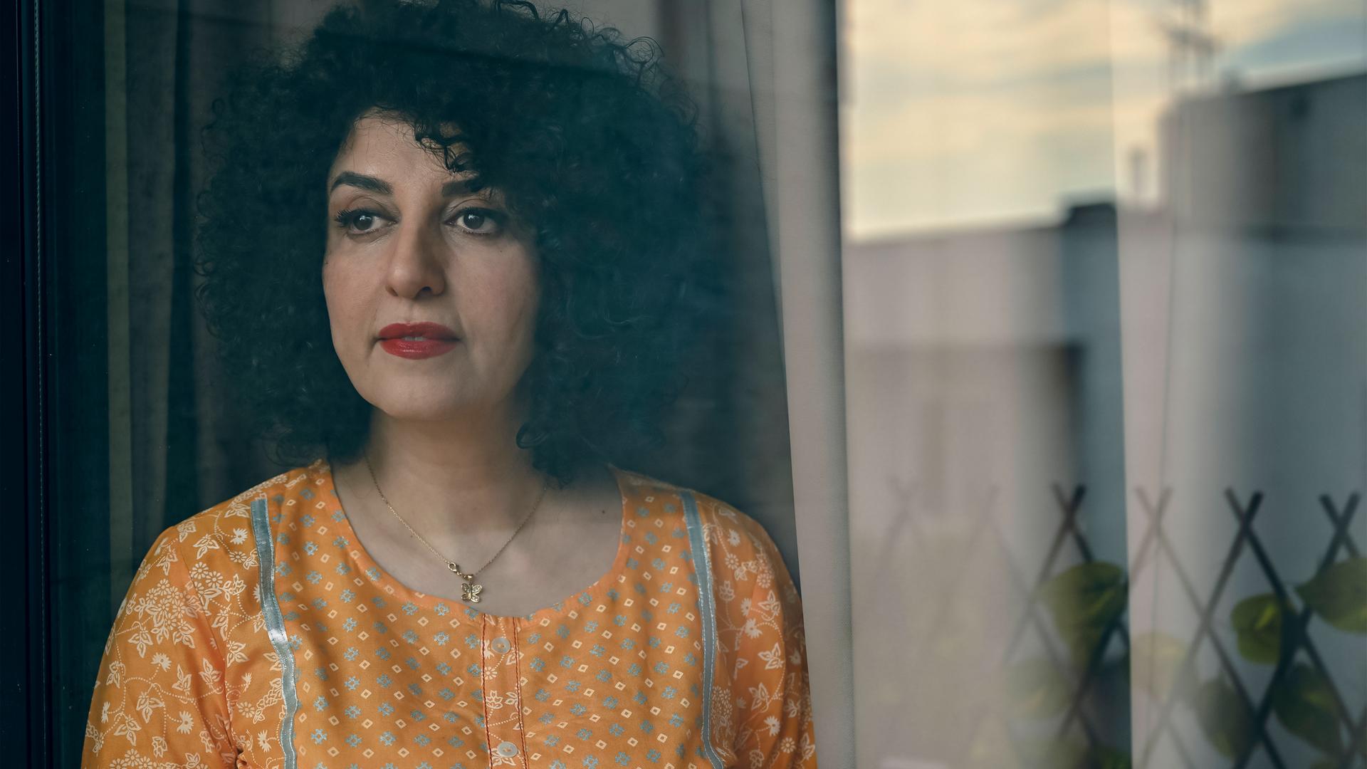 Porträtfoto einer Frau mit dunklem schulterlangem gelocktem Haar, die eine gelbe Bluse trägt und an einem Fenster steht.