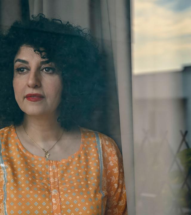 Porträtfoto einer Frau mit dunklem schulterlangem gelocktem Haar, die eine gelbe Bluse trägt und an einem Fenster steht.