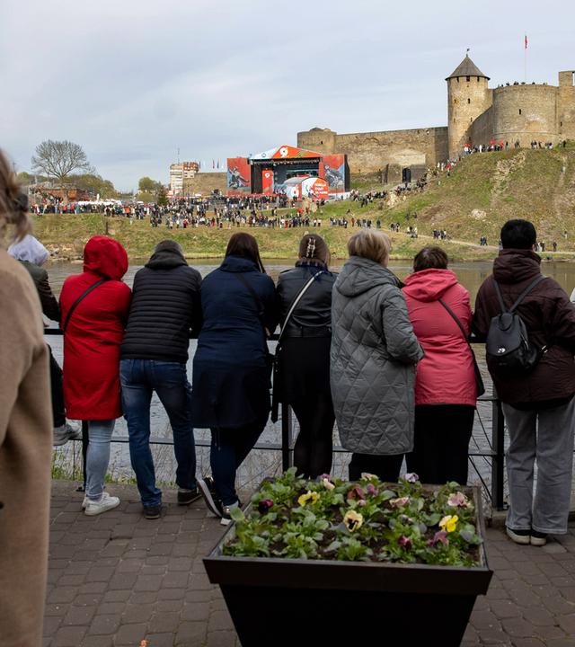 Menschen stehen mit dem Rücken zum Betrachter an einem Geländer und blicken über einen Fluss zum anderen Ufer, wo unterhalb einer Burg eine große Bühne aufgebaut ist, um die sich viele Menschen versammelt haben.