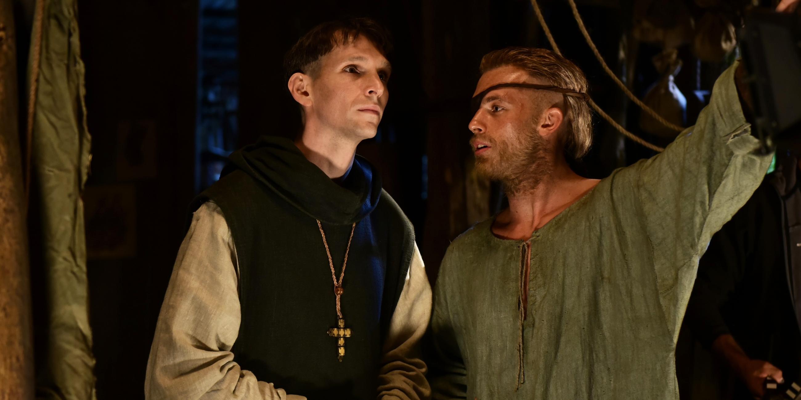Narziss (Sabin Tambrea) und Goldmund (Jannis Niewöhner) stehen nebeneinander im Kloster. Goldmund schaut Narziss an und spricht zu ihm, während er weisend seinen Arm ausstreckt.