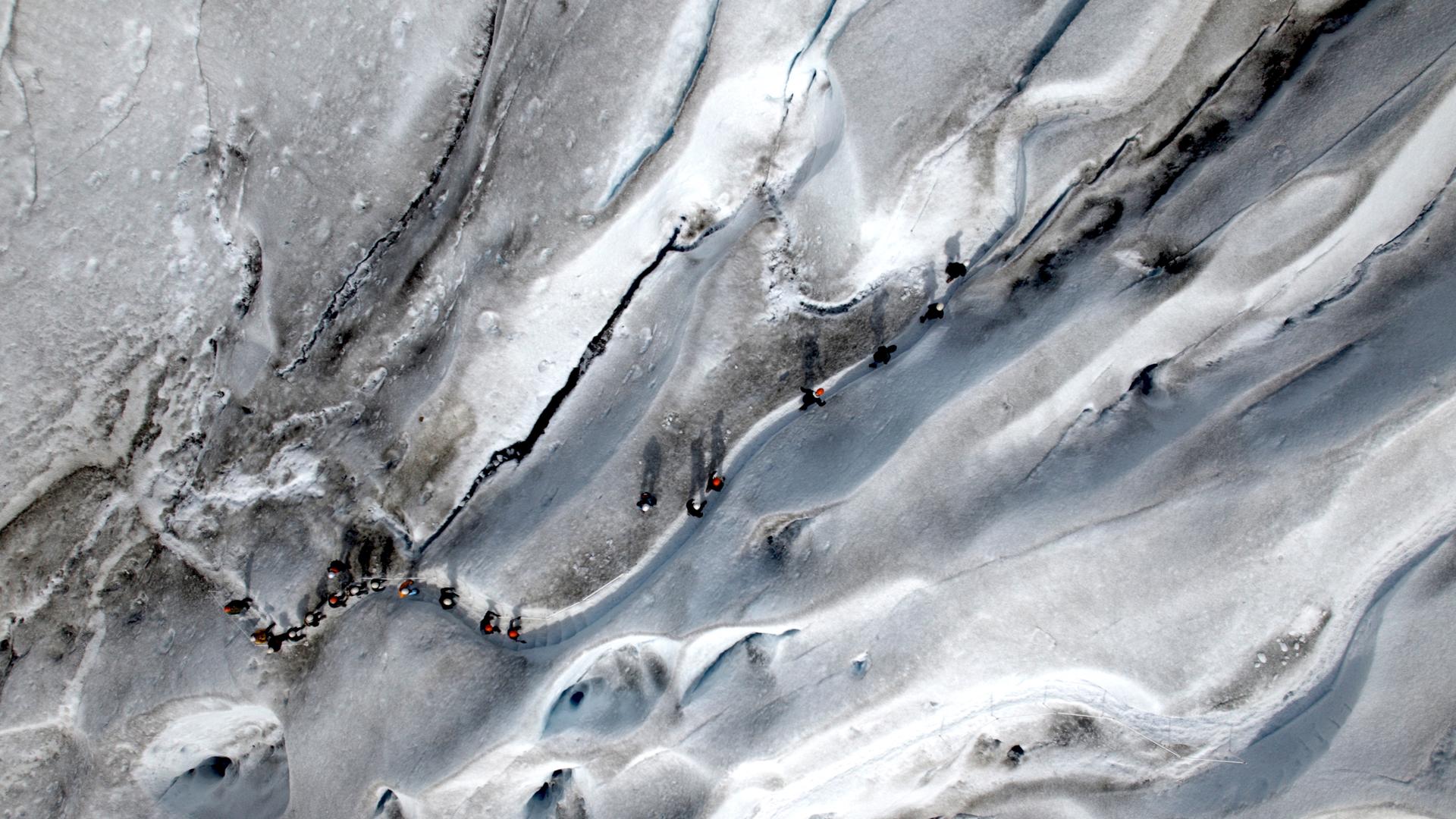 Vogelperspektive: Eine Reihe von Wanderern überquert eine Eisfläche. Das Eis ist stellenweise grau verfärbt