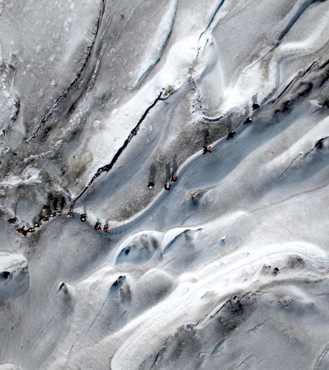 Vogelperspektive: Eine Reihe von Wanderern überquert eine Eisfläche. Das Eis ist stellenweise grau verfärbt