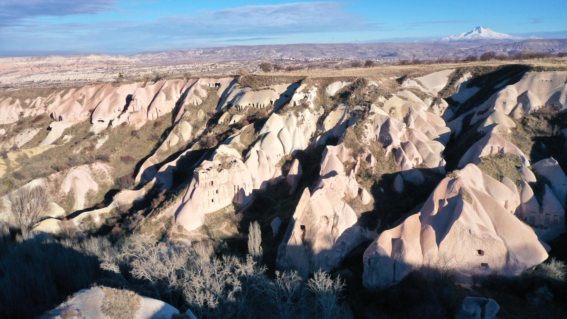  Im Vordergrund befinden sich hellgraue Felswände an einer Talseite, die erkennbare, bearbeitete Öffnungen aufweisen. Dahinter ragt ein weißer Berg in den blauen Himmel.
