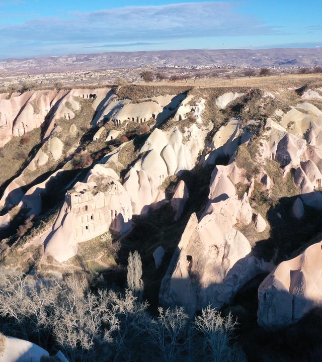  Im Vordergrund befinden sich hellgraue Felswände an einer Talseite, die erkennbare, bearbeitete Öffnungen aufweisen. Dahinter ragt ein weißer Berg in den blauen Himmel.