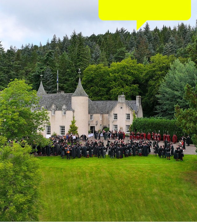 Blick von schräg oben: Vor einem schlossartigen Gebäude in grüner Landschaft stehen Menschen.
