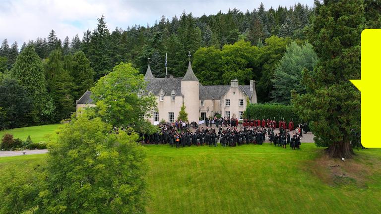  Blick von schräg oben: Vor einem schlossartigen Gebäude in grüner Landschaft stehen Menschen.