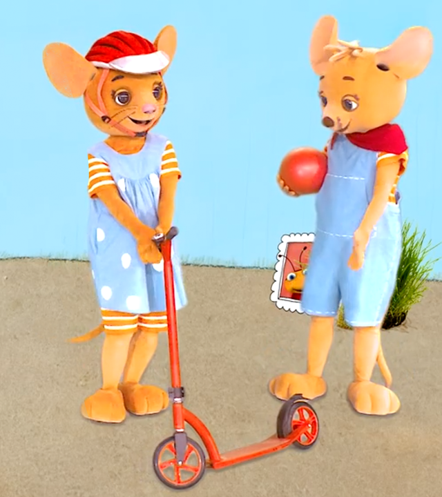 Naya und Jo stehen vor blauem Hintergrund. Naya (links) hat einen roten Roller und trägt einen Schutzhelm. Jo hält einen Ball und zeigt auf den Roller.