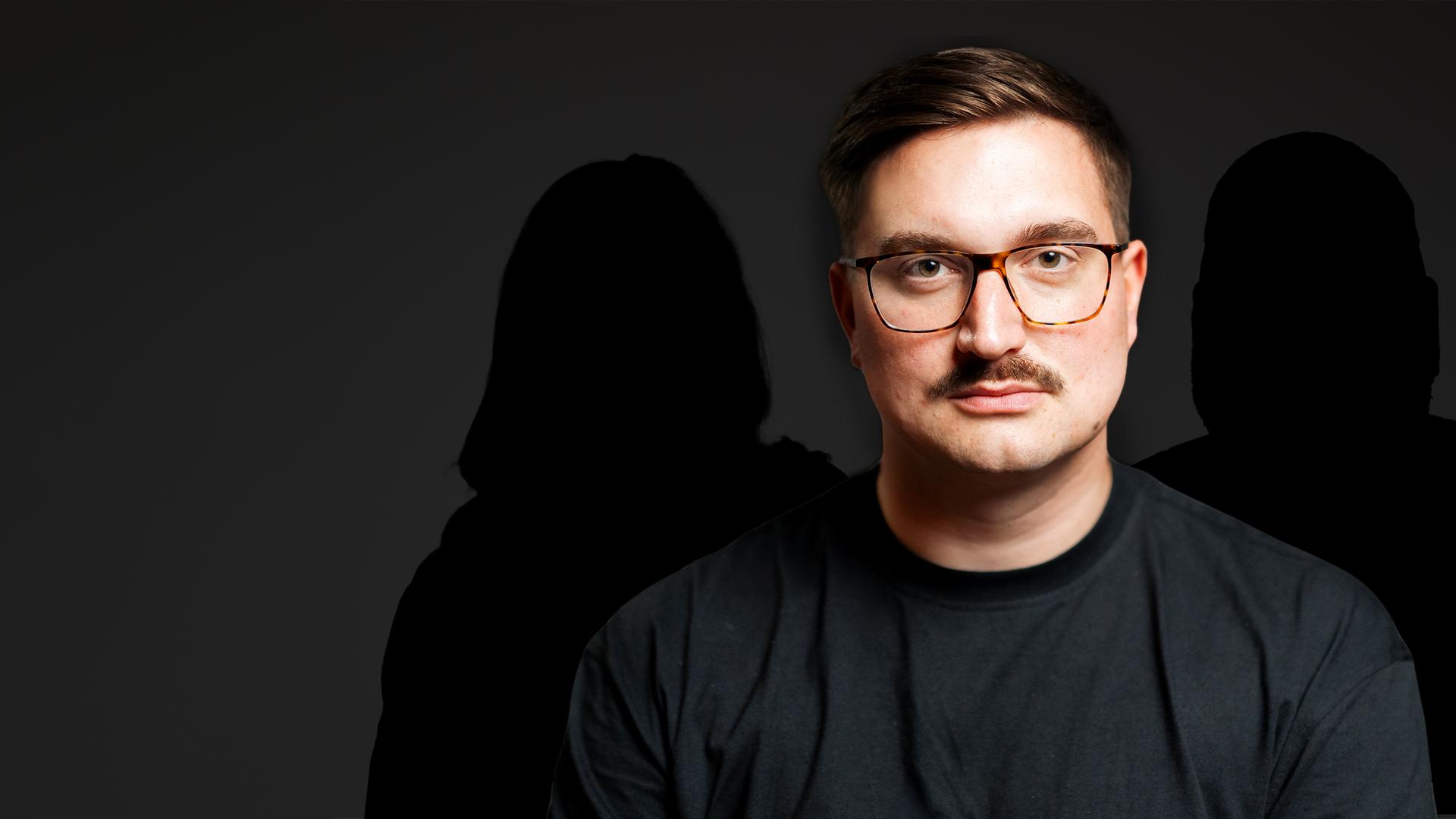 Ein Mann mit Brille und Schnurrbart im Porträt. Hinter ihm sind zwei Schatten zu sehen.