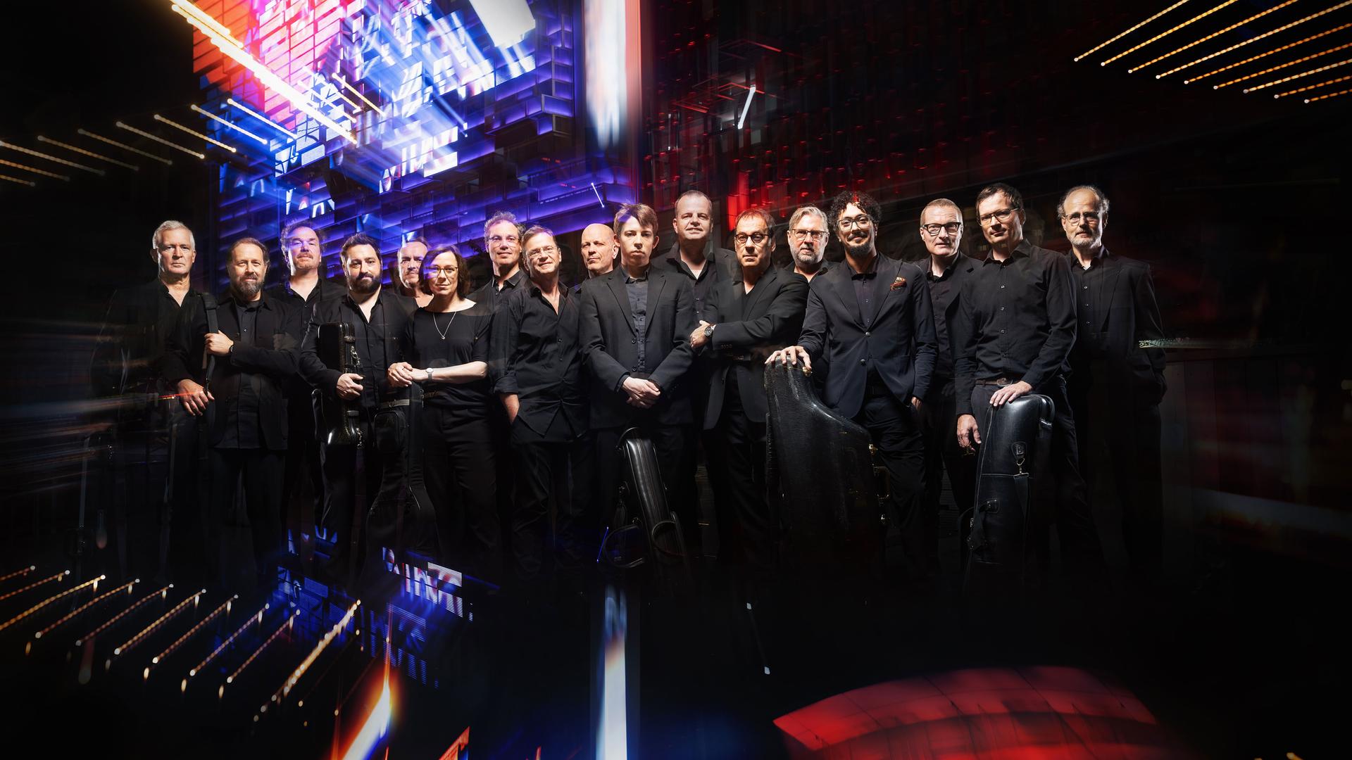 Große Musikgruppe auf einer Bühne mit bunten Lichteffekten; mehrere Musiker mit Instrumenten in Konzertaufstellung.