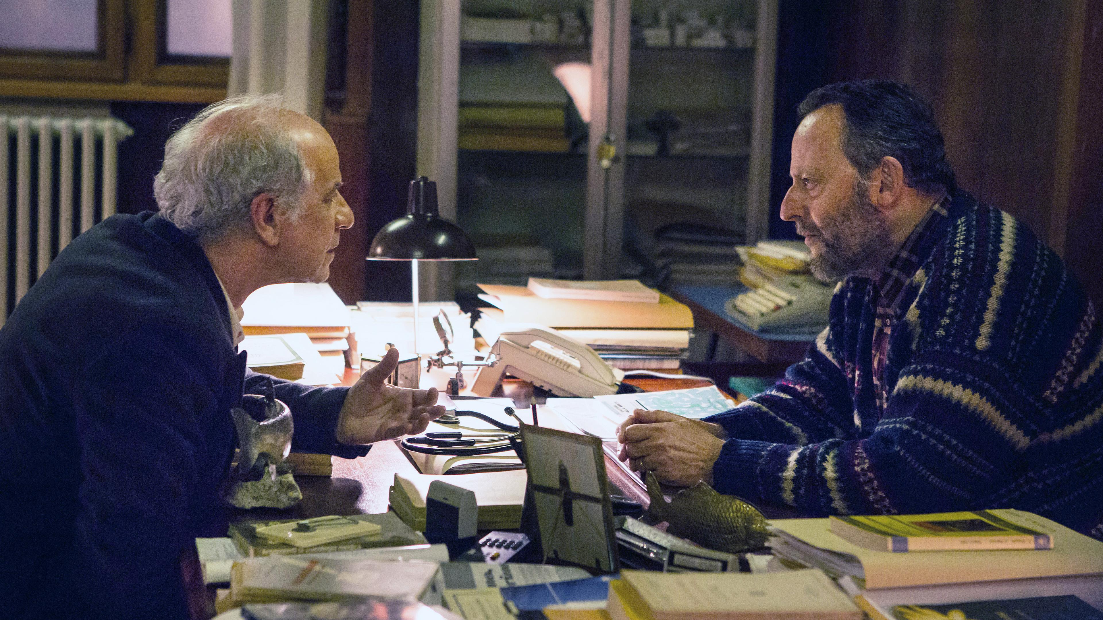 "Der Nebelmann": Vogel (Toni Servillo) und Flores (Jean Reno) sitzen sich an einem mit Büchern und Unterlagen überdeckten Schreibtisch gegenüber und sprechen miteinander.