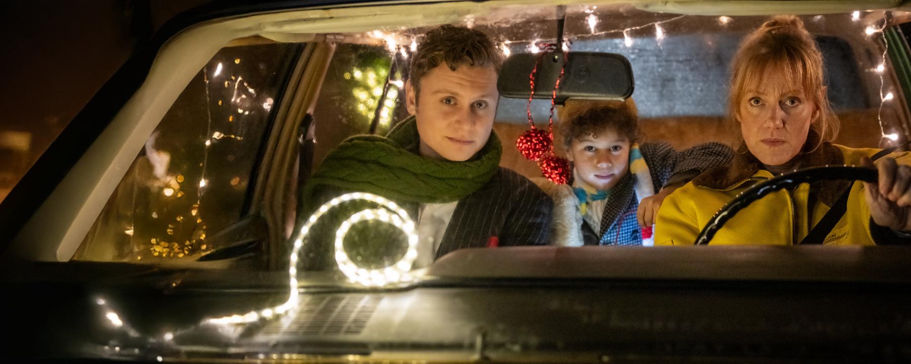 "Nelly und das Weihnachtswunder": Sonny (Rafael Gareisen), Nick (Joah Lion Goetze) und Nelly (Anna Schudt) sitzen in einem Auto, das mit Lichtergirlanden im Inneren geschmückt ist. Nelly sitzt am Steuer, Sonny sitzt neben ihr, Nick sitzt hinter den beiden und hat einen Arm auf Nellys Rückenlehne gelegt. Alle schauen interessiert nach vorne.