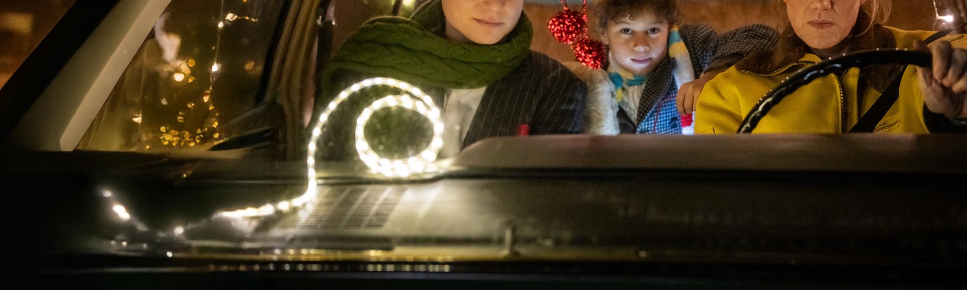 "Nelly und das Weihnachtswunder": Sonny (Rafael Gareisen), Nick (Joah Lion Goetze) und Nelly (Anna Schudt) sitzen in einem Auto, das mit Lichtergirlanden im Inneren geschmückt ist. Nelly sitzt am Steuer, Sonny sitzt neben ihr, Nick sitzt hinter den beiden und hat einen Arm auf Nellys Rückenlehne gelegt. Alle schauen interessiert nach vorne.