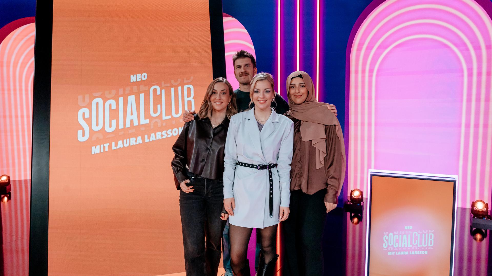 Lena Kupke, Simon Stäblein, Laura Larsson und Maraam (v.l.n.r.) posieren im Studio der Comedy-Gameshow "Neo Social Club". Hinter ihnen steht ein überdimensionierter Smartphone-Bildschirm.