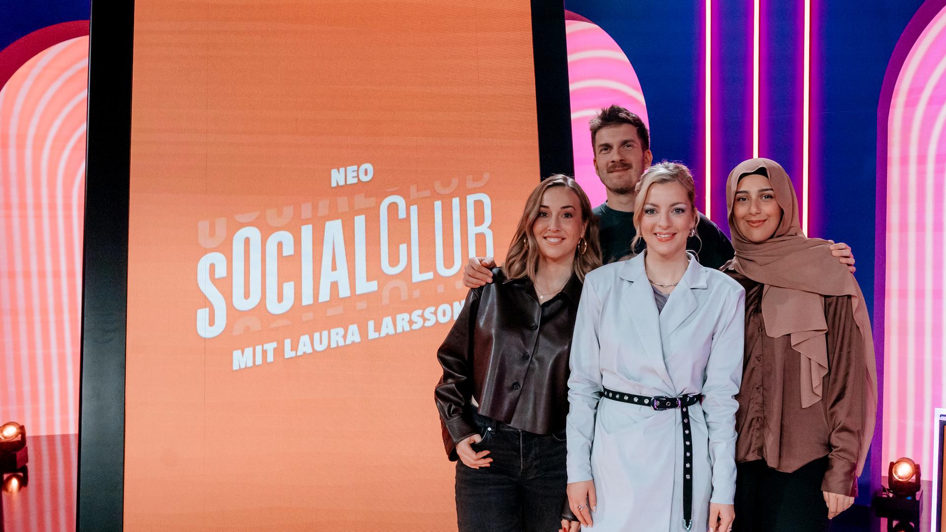 Lena Kupke, Simon Stäblein, Laura Larsson und Maraam (v.l.n.r.) posieren im Studio der Comedy-Gameshow "Neo Social Club". Hinter ihnen steht ein überdimensionierter Smartphone-Bildschirm.