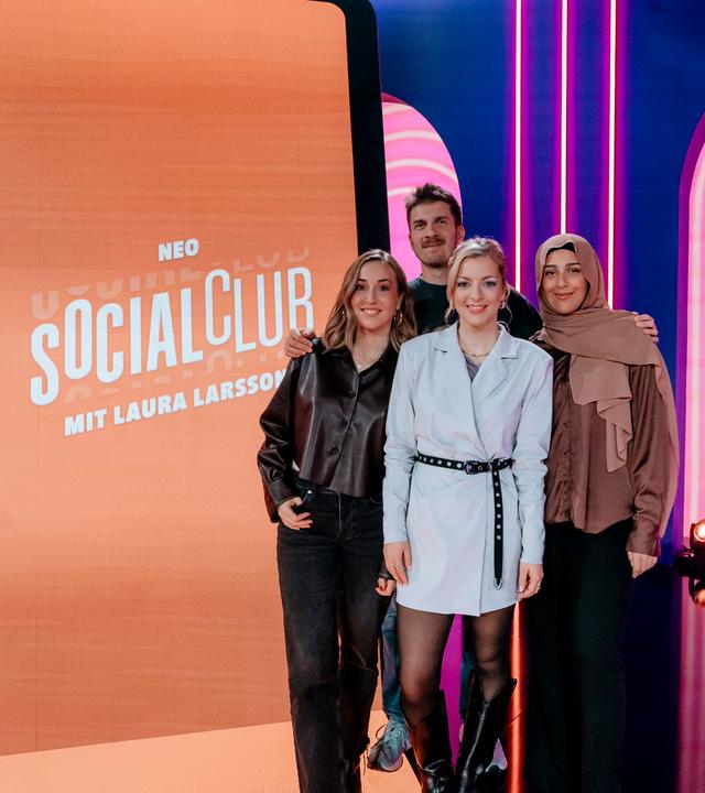 Lena Kupke, Simon Stäblein, Laura Larsson und Maraam (v.l.n.r.) posieren im Studio der Comedy-Gameshow "Neo Social Club". Hinter ihnen steht ein überdimensionierter Smartphone-Bildschirm.