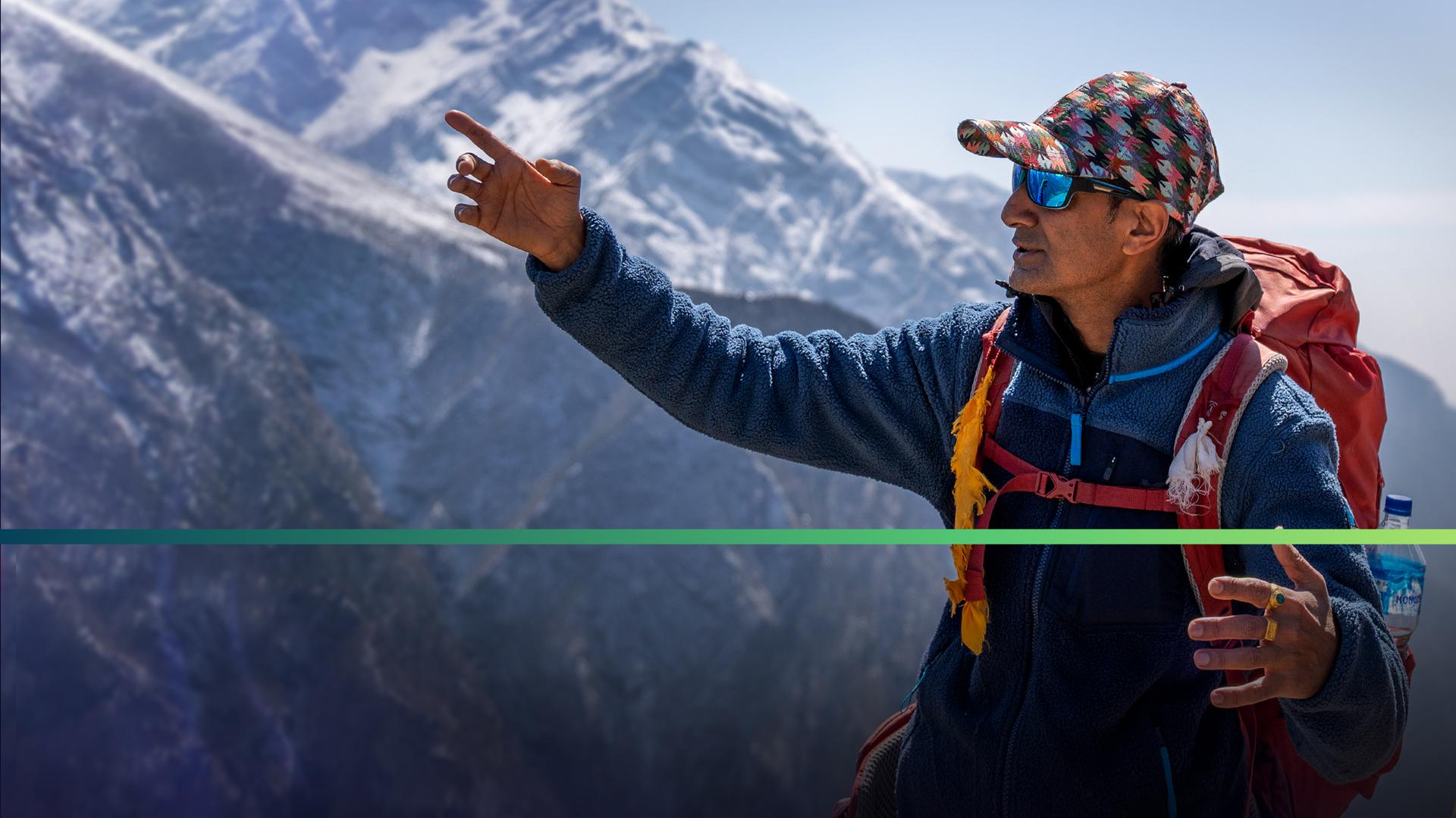 Zwei Männer in Bergsteiger-Kleidung stehen vor dem Hintergrund eines Bergmassivs. Der linke Mann deutet nach oben, der Blick des rechten folgt lächelnd der Hand.