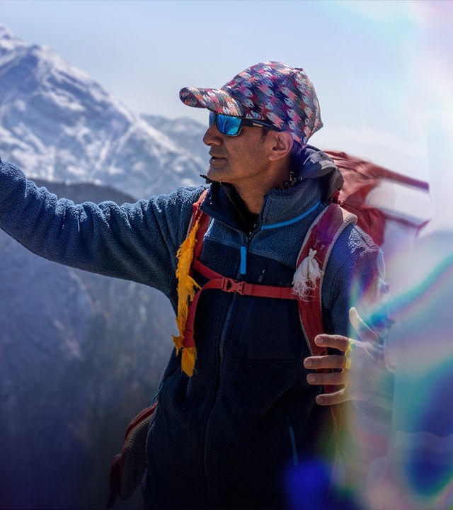 Zwei Männer in Bergsteiger-Kleidung stehen vor dem Hintergrund eines Bergmassivs. Der linke Mann deutet nach oben, der Blick des rechten folgt lächelnd der Hand.
