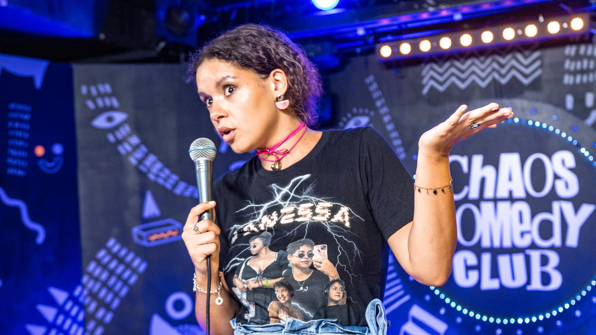 Vanessa Ebert steht in der Mitte des Bildes. In der rechten Hand hält sie ein Mikrofon. Ihren linken Arm streckt die angewinkelt zur rechten Bildseite. 