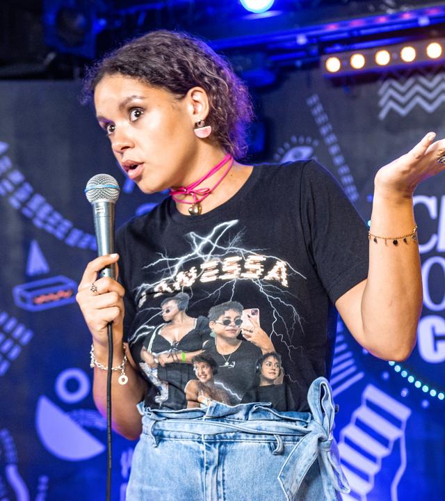 Vanessa Ebert steht in der Mitte des Bildes. In der rechten Hand hält sie ein Mikrofon. Ihren linken Arm streckt die angewinkelt zur rechten Bildseite. 