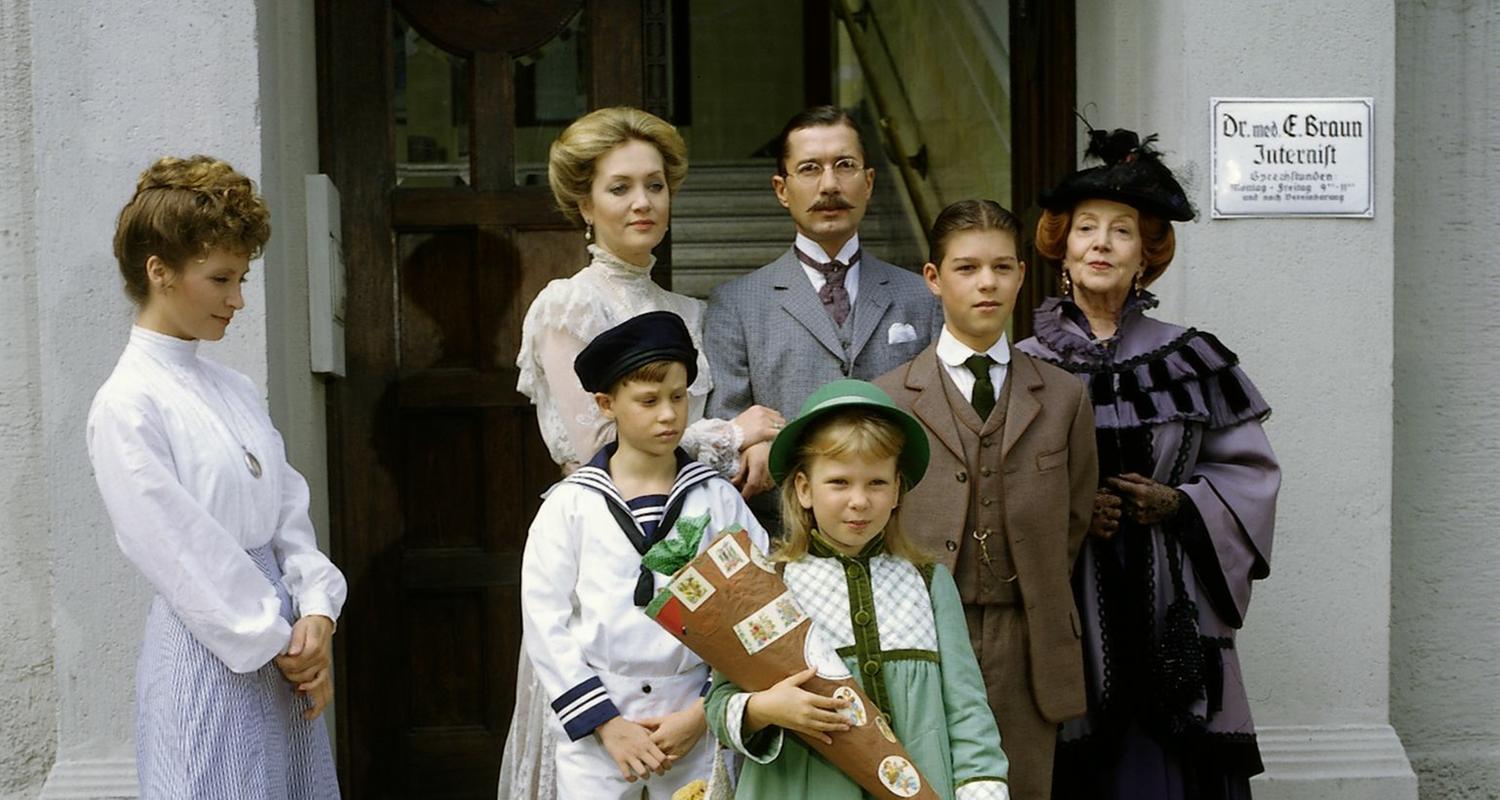 "Das Nesthäkchen - Teil 3": Annemarie (Kathrin Toboll) steht mit einer Schultüte vor der Schule. Hinter ihr stehen Dr. Ernst Braun (Christian Wolff), Tante Käthe (Dagmar Hessenland), Onkel Heinrich (Willy Harlander), Elsbeth (Doris Kunstmann), Frl. Hering (Wega Jahnke) sowie ihre Brüder Klaus (Florian Baier) und Hans (Oliver Schlicht).