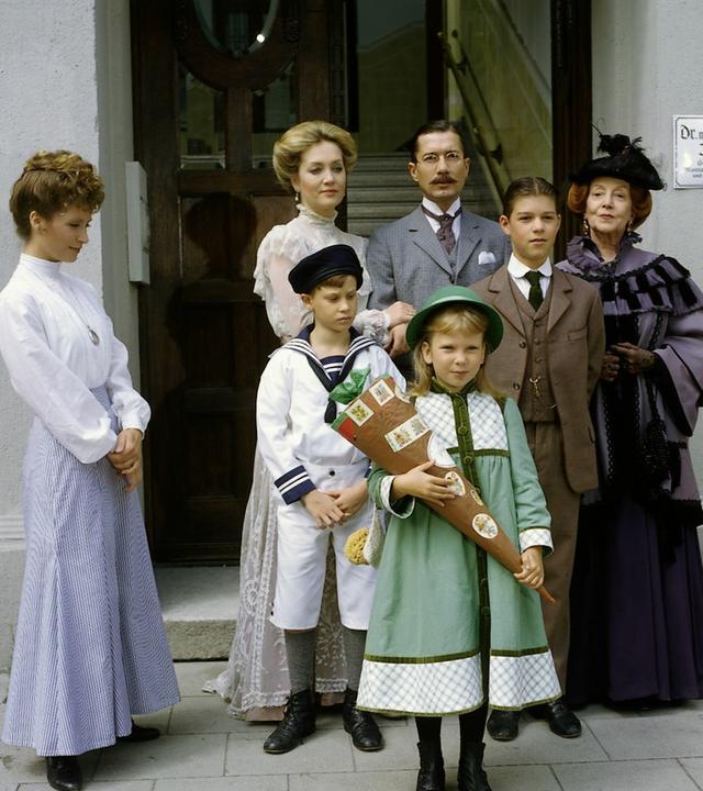 "Das Nesthäkchen - Teil 3": Annemarie (Kathrin Toboll) steht mit einer Schultüte vor der Schule. Hinter ihr stehen Dr. Ernst Braun (Christian Wolff), Tante Käthe (Dagmar Hessenland), Onkel Heinrich (Willy Harlander), Elsbeth (Doris Kunstmann), Frl. Hering (Wega Jahnke) sowie ihre Brüder Klaus (Florian Baier) und Hans (Oliver Schlicht).