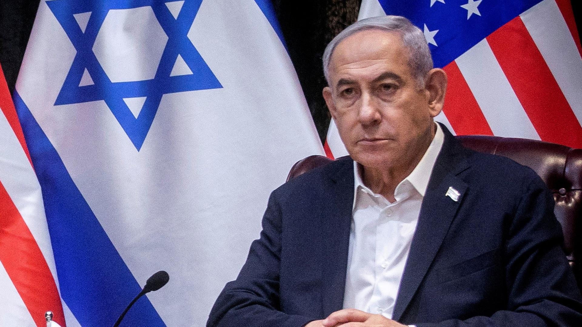 Benjamin Netanjahu, ein älterer Mann mit grauen Haaren, sitzt vor einer israelischen und einer US-amerikanischen Flagge in einer Pressekonferenz.