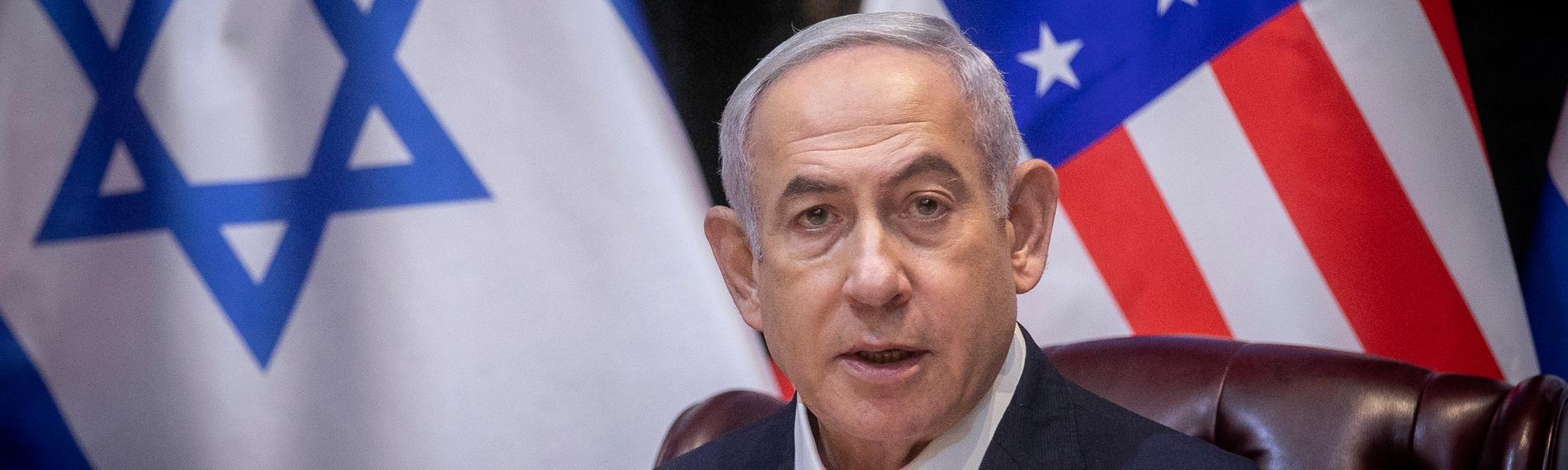 Benjamin Netanjahu, ein älterer Mann mit grauen Haaren, sitzt vor einer israelischen und einer US-amerikanischen Flagge in einer Pressekonferenz.