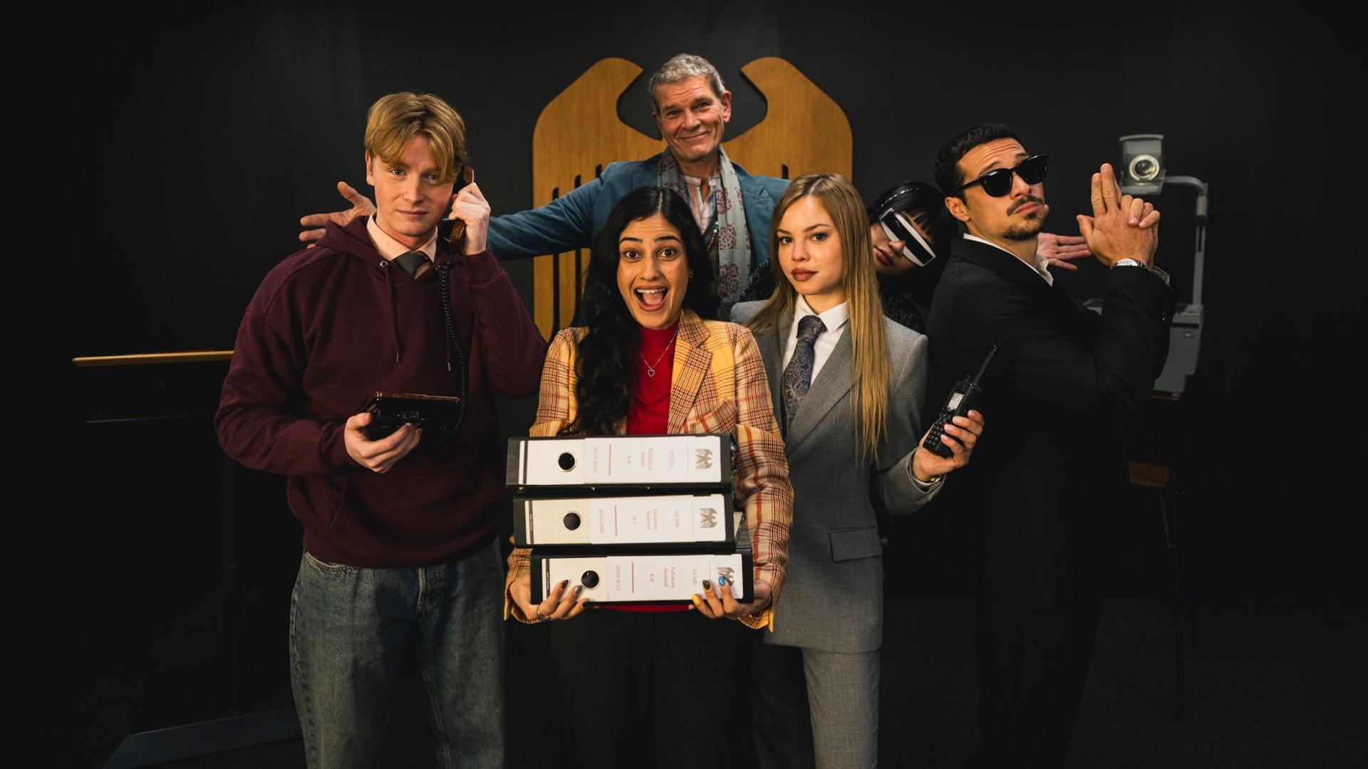 Sechs Spione stehen in lustigen Posen vor einer Wand, im Hintergrund ist der Bundesadler zu sehen.