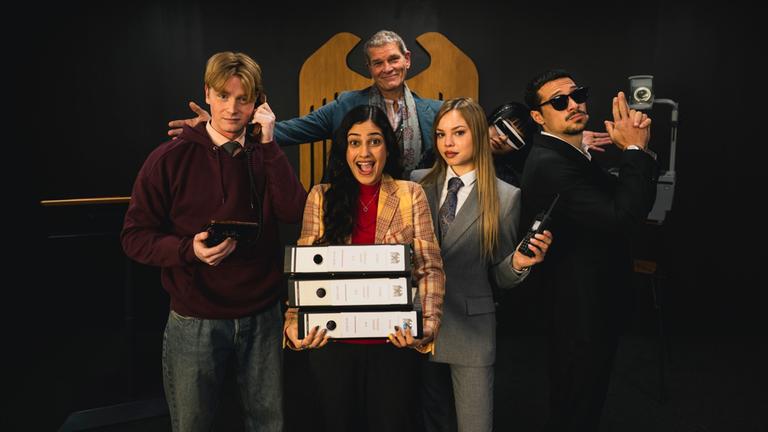 Sechs Spione stehen in lustigen Posen vor einer Wand, im Hintergrund ist der Bundesadler zu sehen.