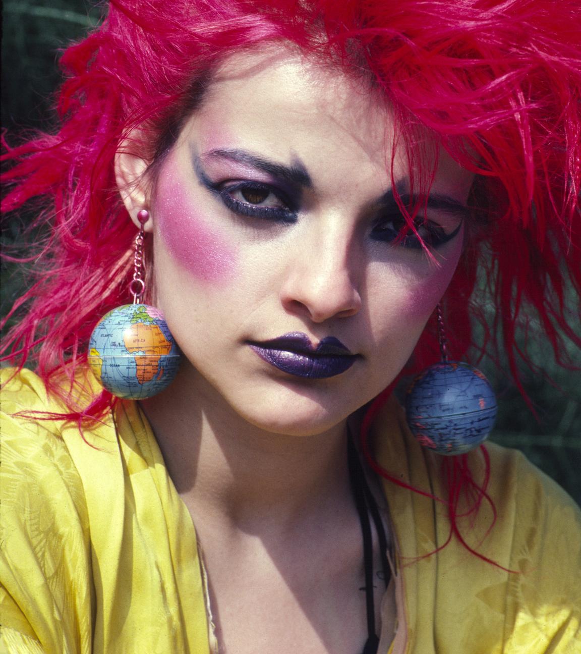 Eine buntgeschmickte Frau mit pinken hochgegelten Haaren und großen Globus-Ohrringen schaut direkt in die Kamera. 