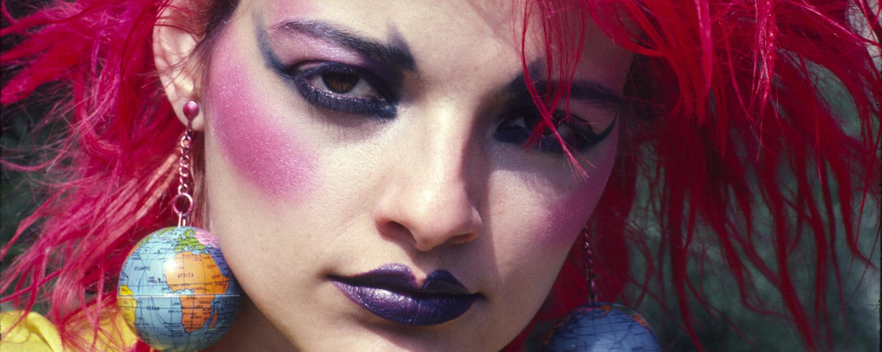 Eine buntgeschmickte Frau mit pinken hochgegelten Haaren und großen Globus-Ohrringen schaut direkt in die Kamera. 