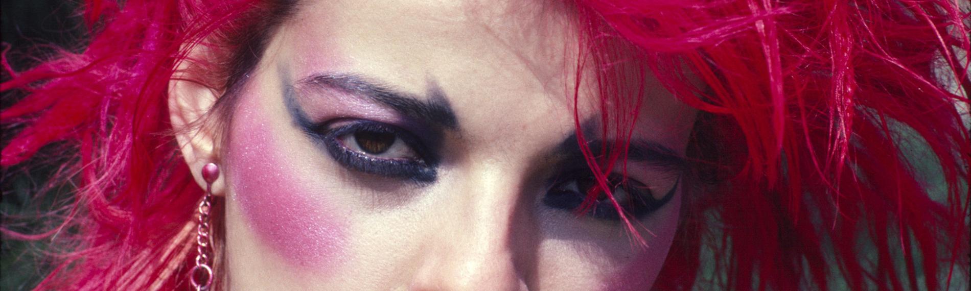 Eine buntgeschmickte Frau mit pinken hochgegelten Haaren und großen Globus-Ohrringen schaut direkt in die Kamera. 