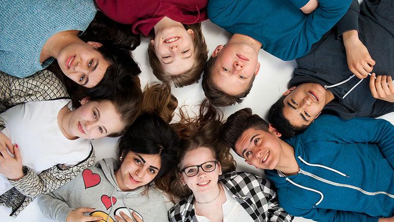 Berlin und wir!: Die acht Protagonisten blicken, sternförmig mit den Köpfen zusammen auf dem Boden liegend, in die Kamera