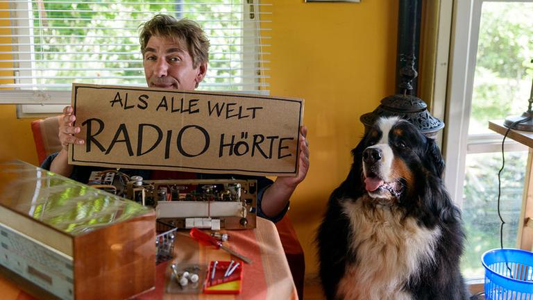 Fritz Fuch sitzt im Bauwagen und hält ein Pappschild hoch auf dem steht "ALS ALLE WELT RADIO HÖRTE". Keks sitzt daneben.