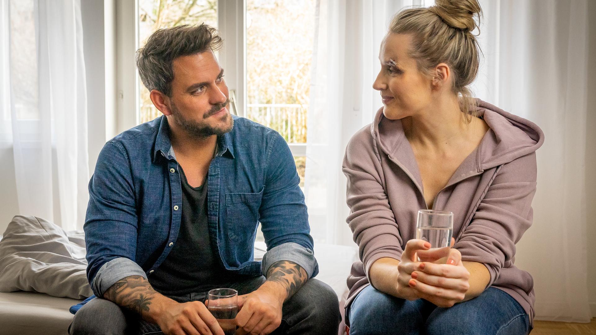 "Notruf Hafenkante - Neues Spiel, neues Glück": Kris (Marc Barthel) und Lena (Anna Hofbauer) sitzen nebeneinander auf einer Bettkante und lächeln sich an. Beide halten ein Glas Wasser in den Händen.